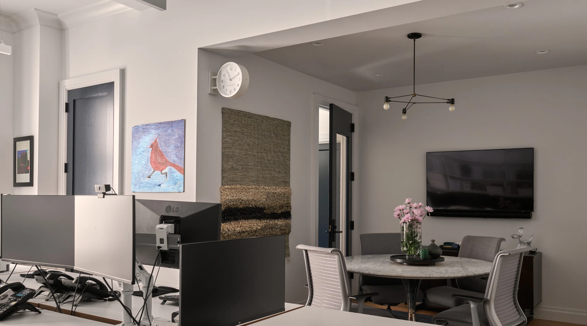 Modern office space with desks, monitors, a round table with chairs, a wall TV, and floral centerpiece.