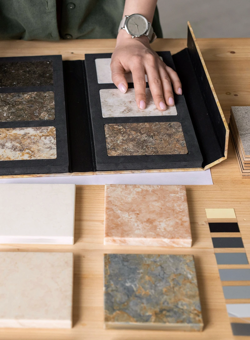 Person's hand touching tile samples in a folder with various tiles and color swatches on wooden table.