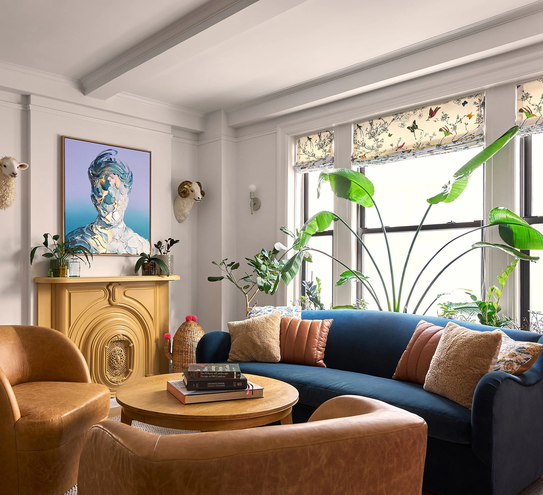 Cozy living room with blue sofa, tan leather chairs, wooden table, plants, and abstract artwork above fireplace.