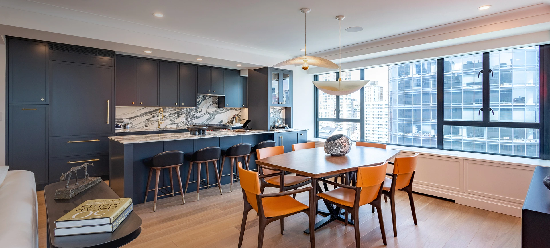 Modern kitchen and dining area with navy cabinets, marble backsplash, wooden table, and orange chairs.