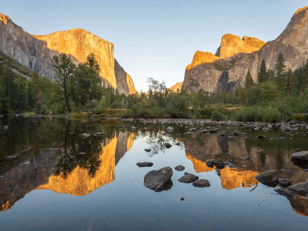 Sunlit mountain cliffs reflected in a calm river with rocks and green trees along the shore.