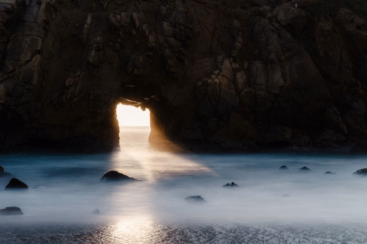 Sunlight shining through a rocky sea cave arch onto misty ocean waters with scattered rocks.