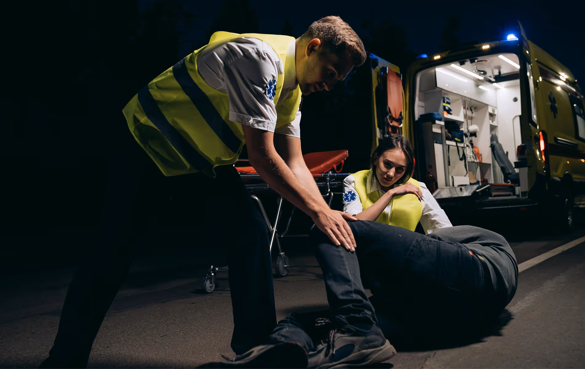 Two paramedics in reflective vests attending to a person lying on the road next to an ambulance at night.