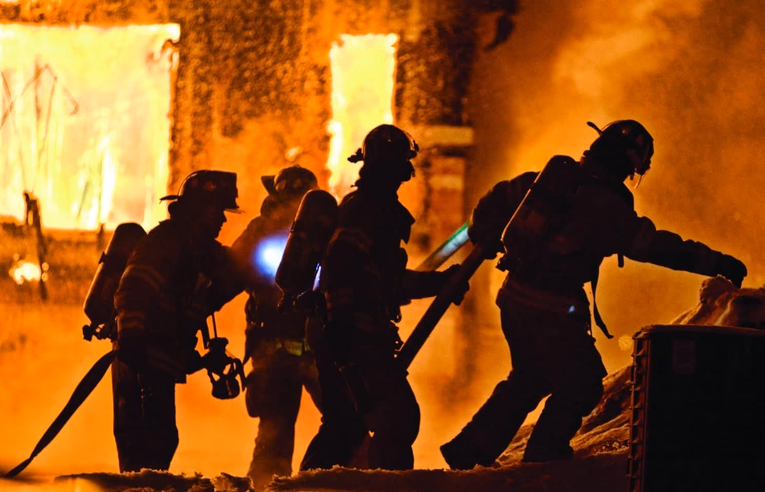Silhouettes of Edmonton Firefighters battling a large blaze at night with flames in the background.