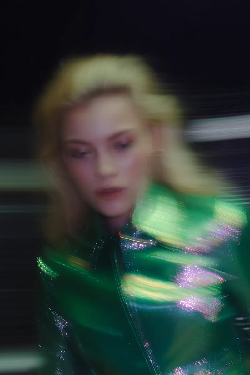 Blurred portrait of a person with blonde hair wearing a shiny green jacket against a dark background.