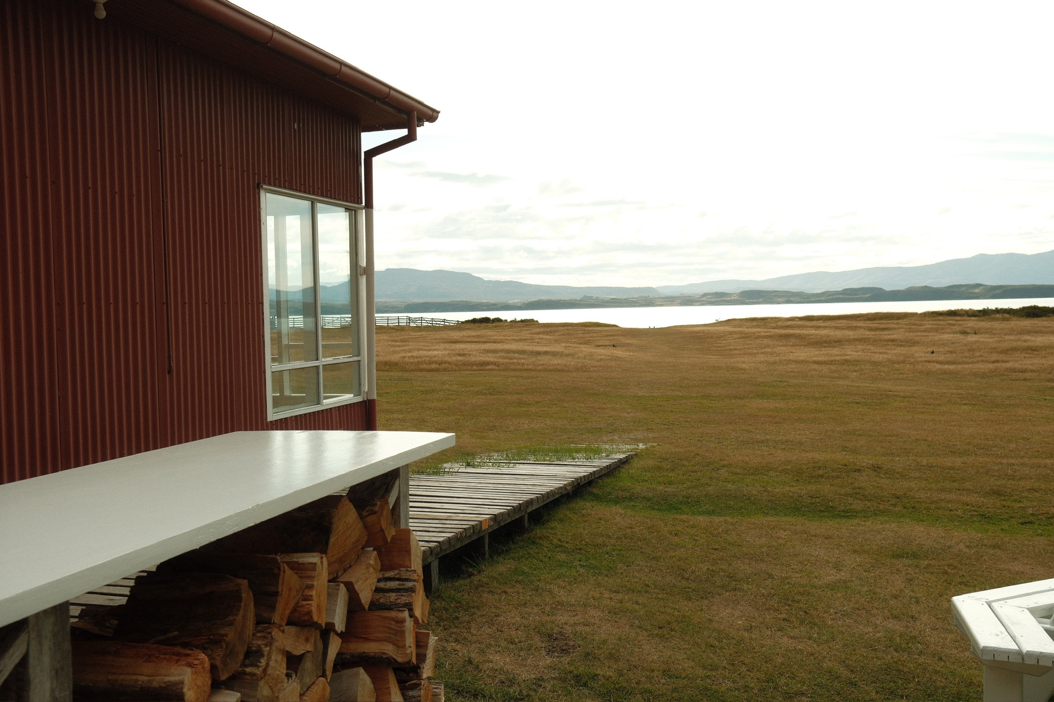 Red corrugated ranch building with stacked firewood overlooking grassland and water