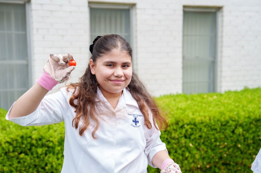 Student in uniform holding a small tomato while gardening outdoors