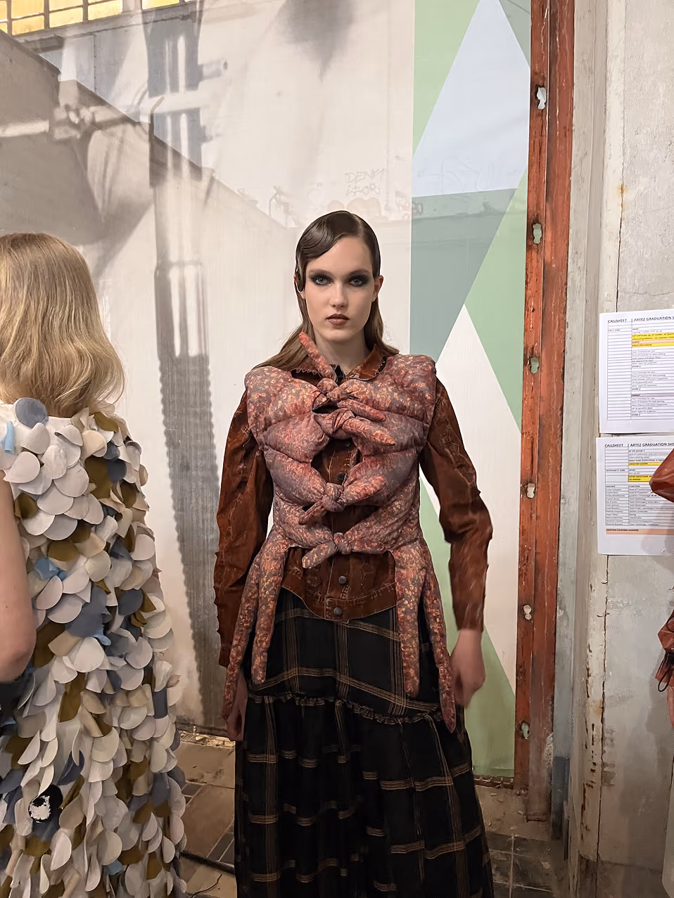 Ein weibliches Model trägt eine braune, strukturierte Jacke mit großen, gebundenen Stoffdetails und einen schwarzen Rock mit Karomuster.