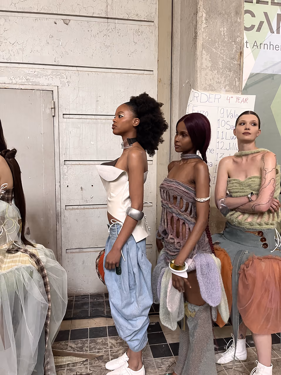 Drei Models stehen in einer Reihe und tragen avantgardistische Outfits in einer industriellen Kulisse.
