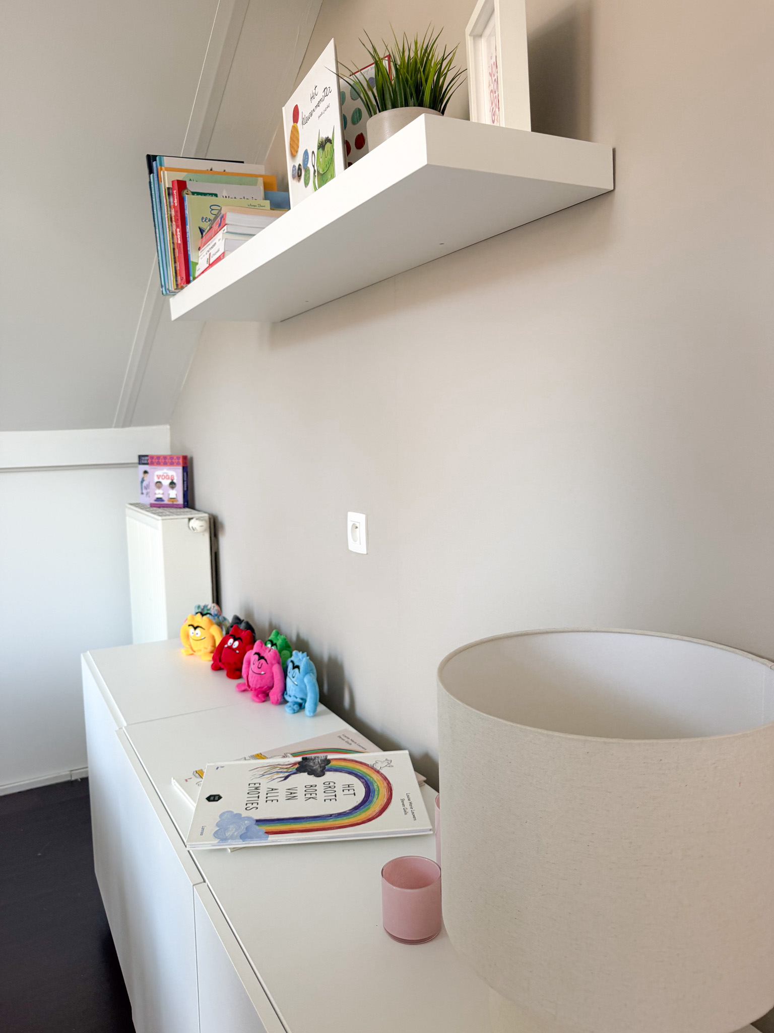 White shelf and cabinet with books, colorful plush toys, a lamp, and a pink candle on a beige wall.