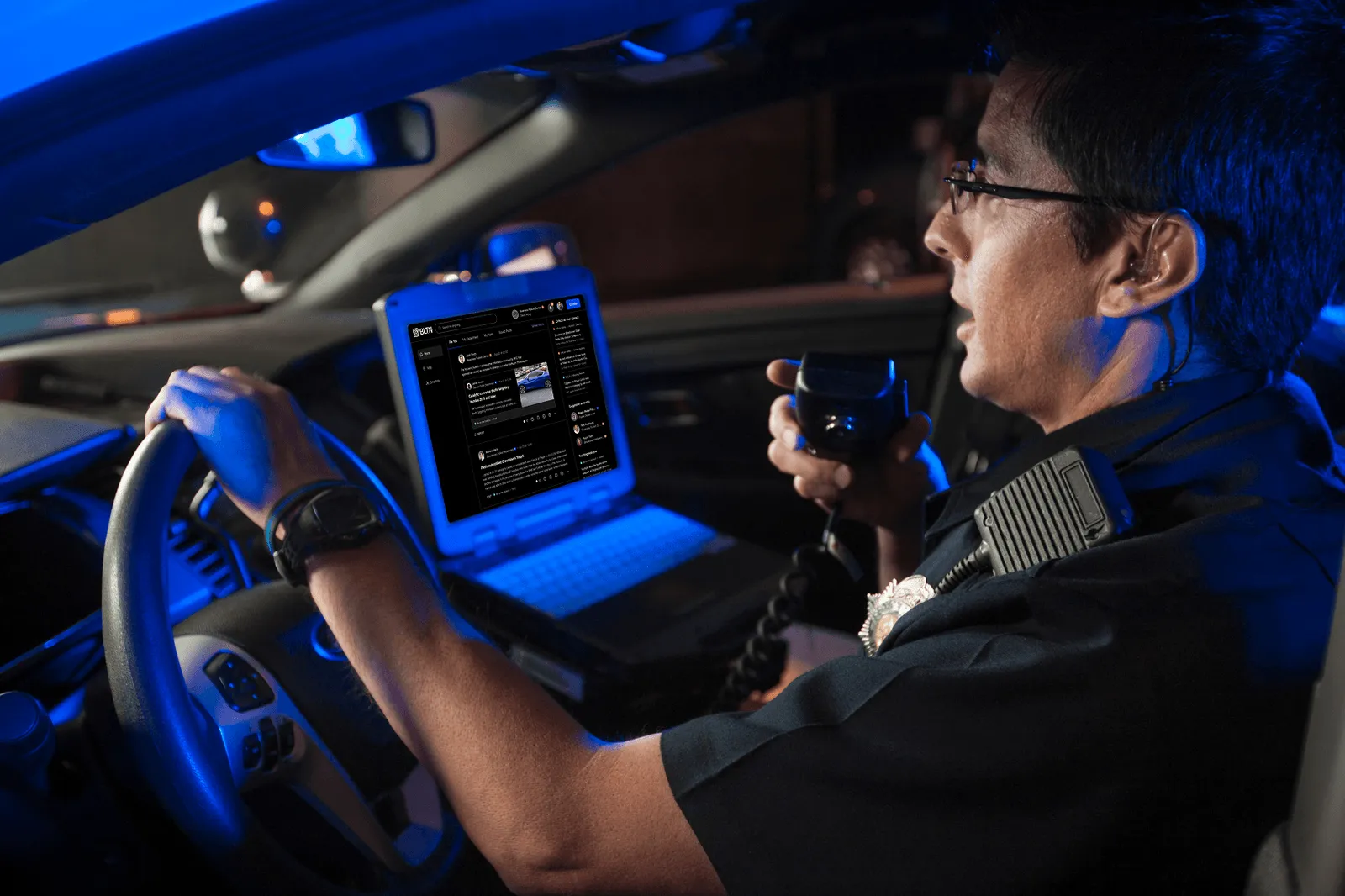 Police officer in car holding radio microphone with open laptop showing social media screen.