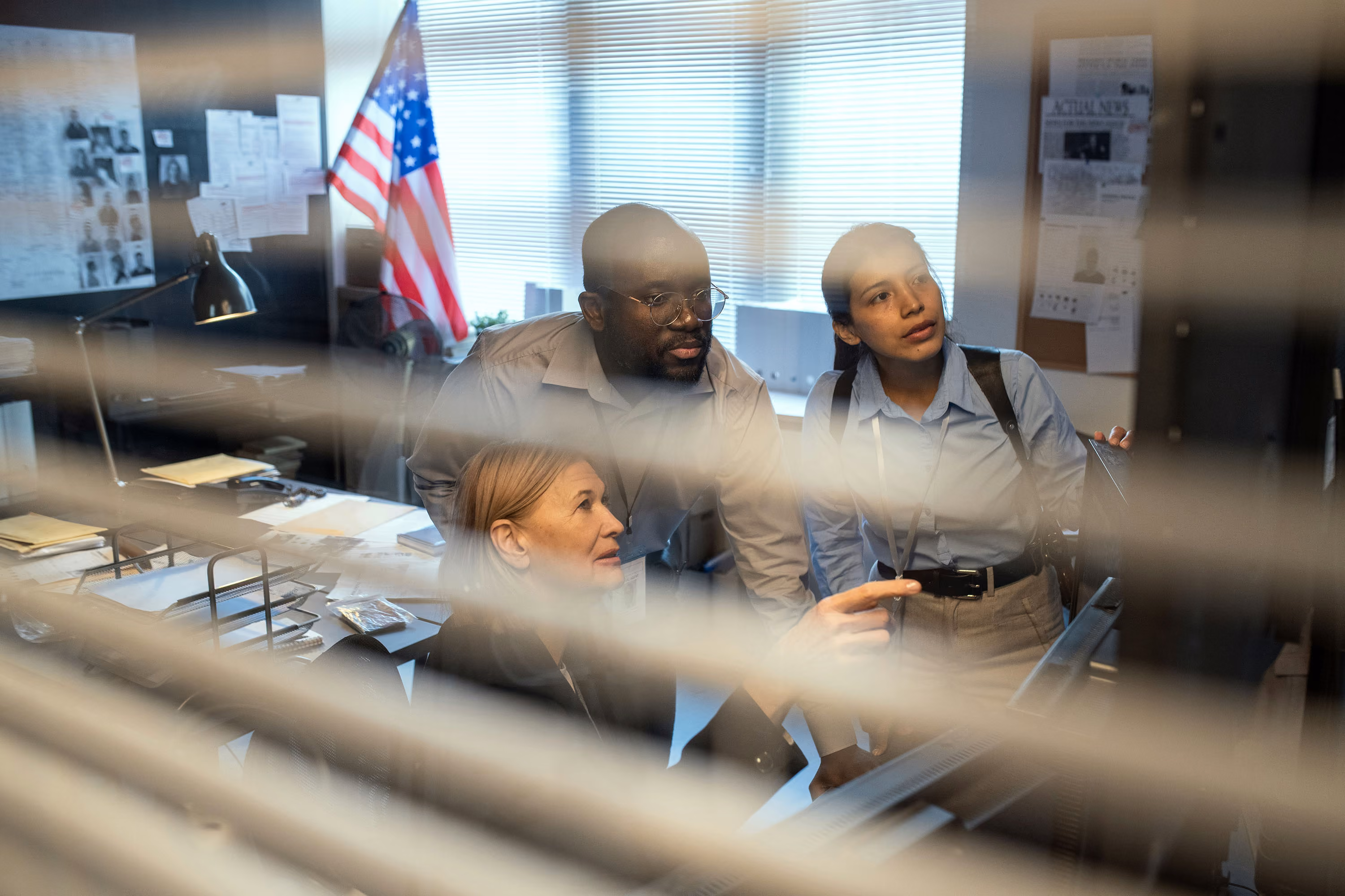 Three coworkers collaborate intently in an office, looking at a computer monitor behind window blinds.