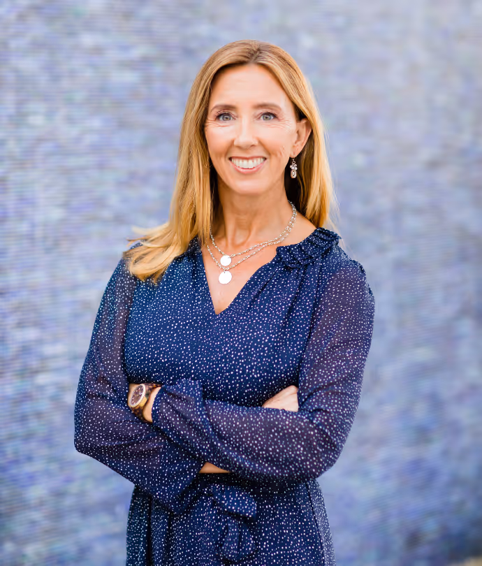 Smiling woman with long blonde hair wearing a navy polka dot dress posing with arms crossed.