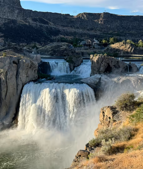 Wide waterfall cascading down rocky cliffs with mist and shrubs in foreground and houses in background.