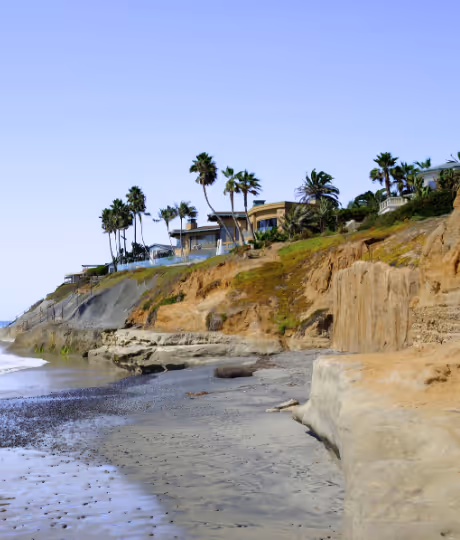 Cliffside houses and palm trees overlooking a sandy beach under a clear blue sky.