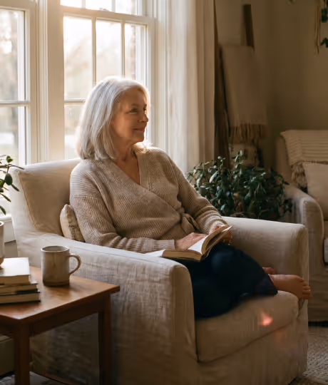 A woman with gray hair sits in a beige armchair reading a book by a large sunlit window.