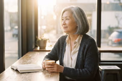Older woman sitting in cafe by window, holding coffee cup, with an open book on table.
