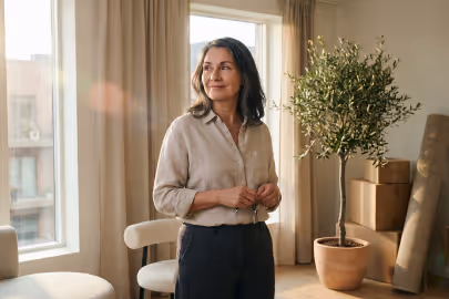 Smiling woman standing in a sunlit room with plants and cardboard boxes.