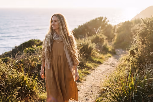 Woman with long blonde hair in brown dress and scarf walking along a coastal trail at sunset.