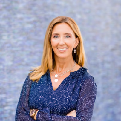 Smiling woman with long blonde hair in a blue patterned blouse standing with arms crossed.