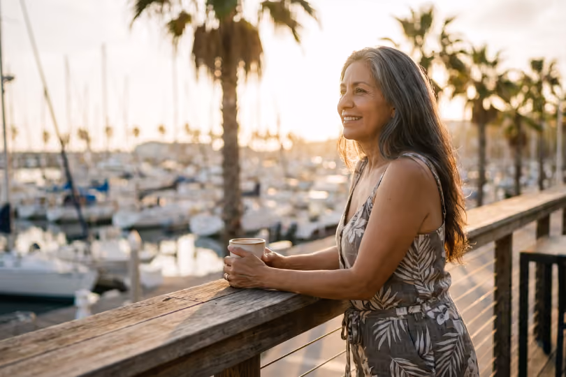 Smiling woman holding a cup while watching boats at a marina during sunset.