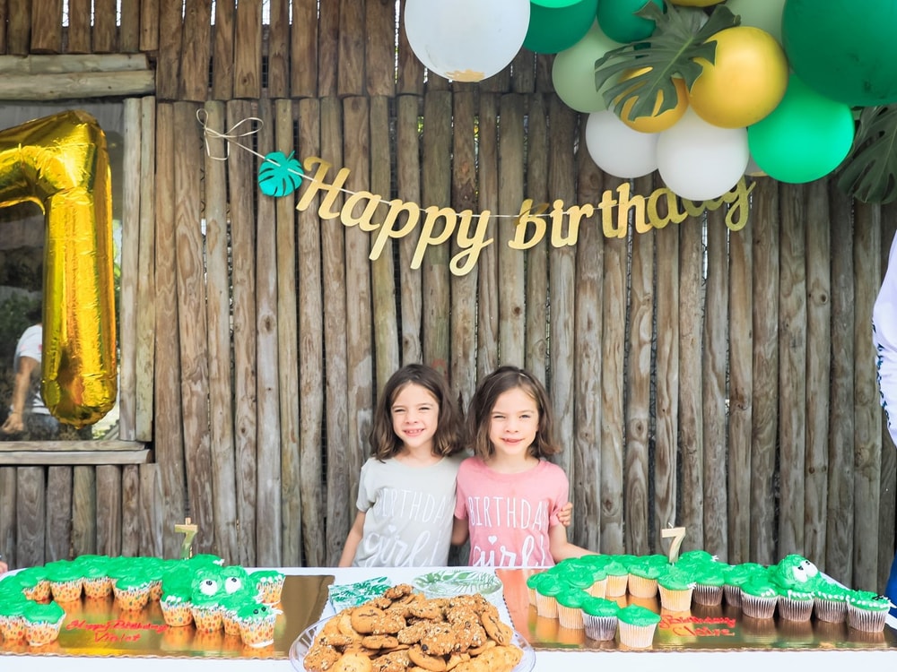 Two girls smiling behind a birthday table of alligator cupcakes and cookies under a Happy Birthday banner at Everglades Alligator Farm