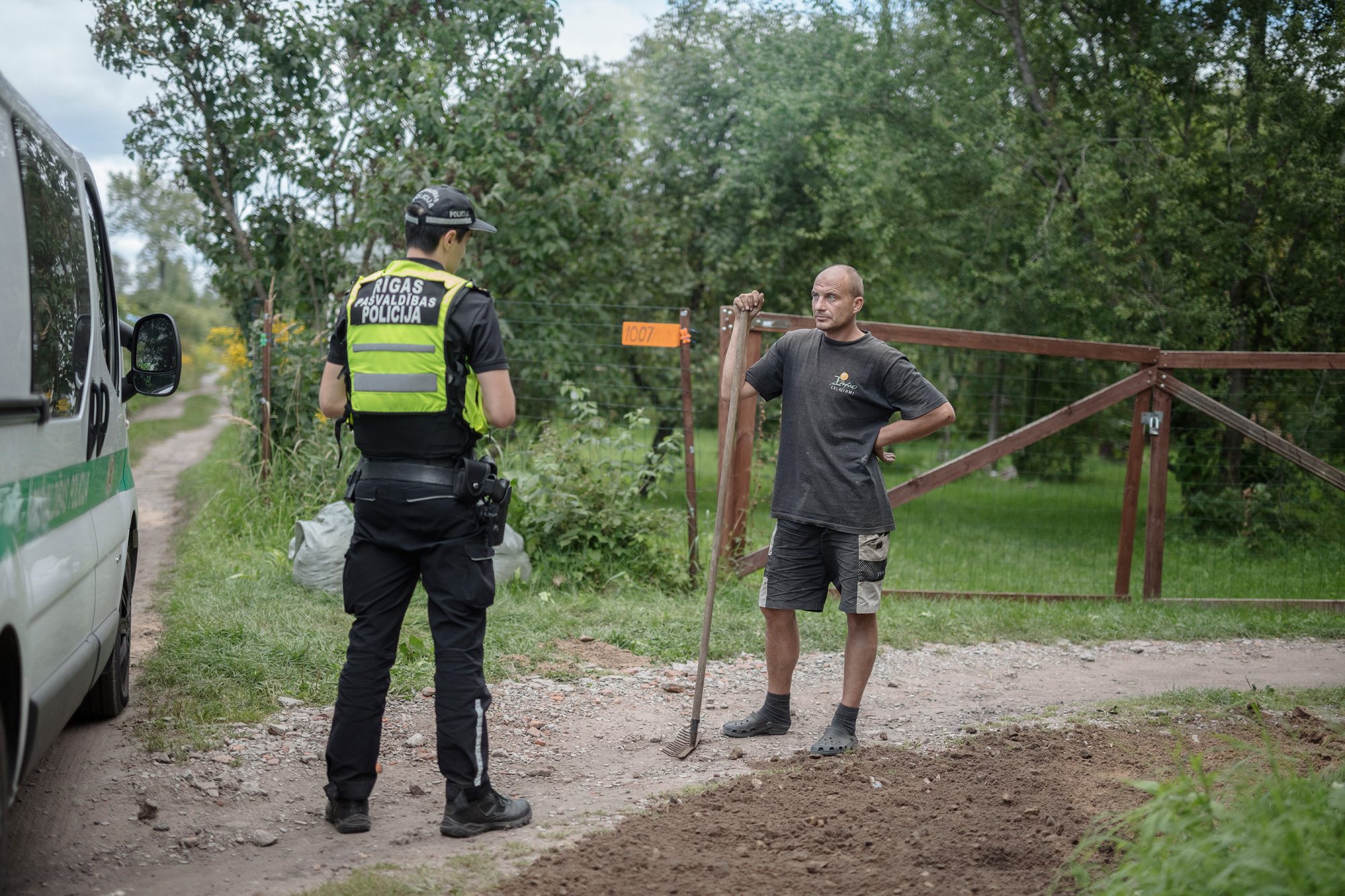 Rīgas pašvaldības policijas darbinieks sarunājas ar vīrieti, kas ar grābekli stāv uz svaigi uzbērtas grants dārziņu ceļā