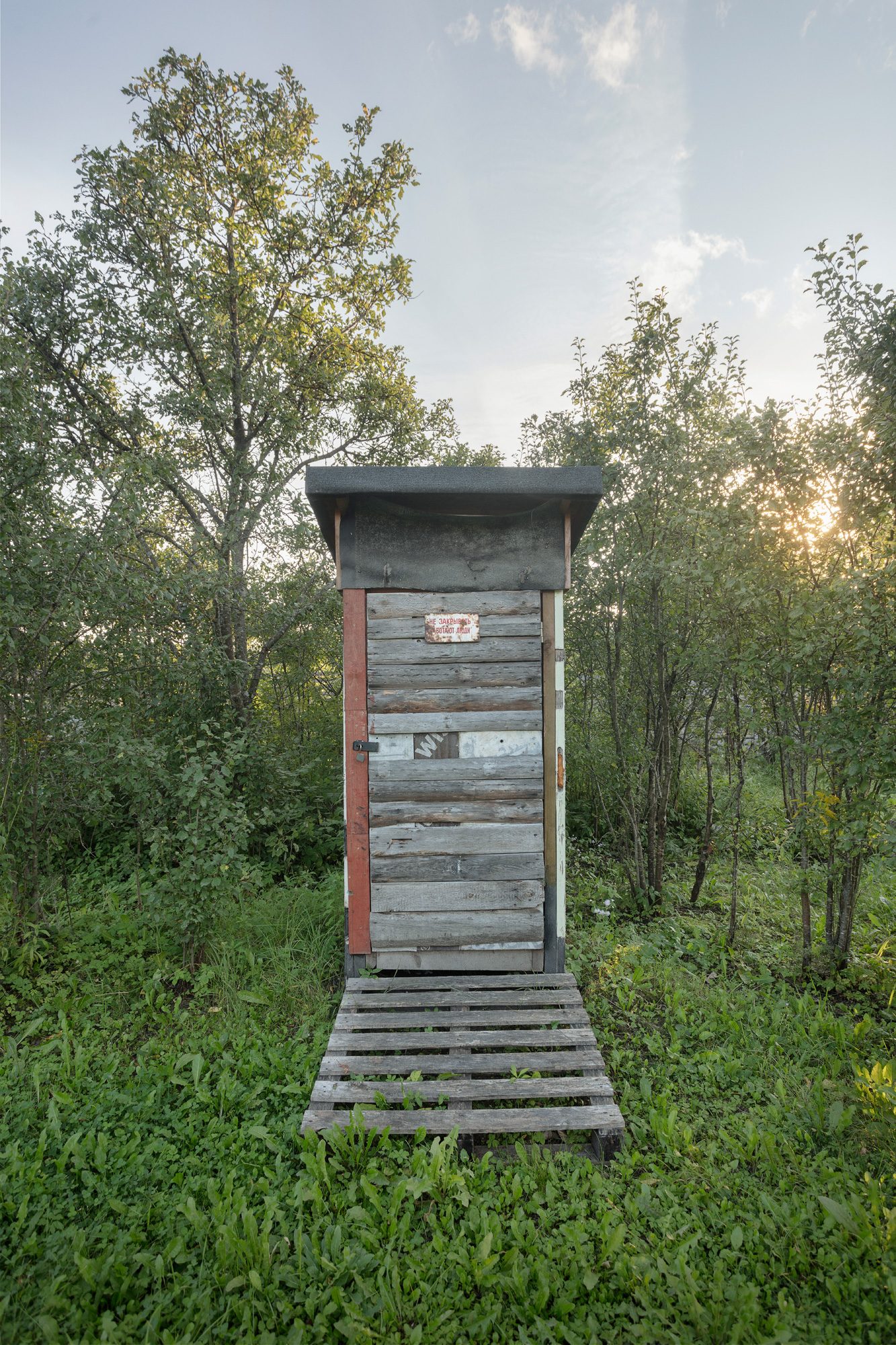 Tumša koka āra tualete uz paliktņiem zālē, aiz tās vakara saule kokos