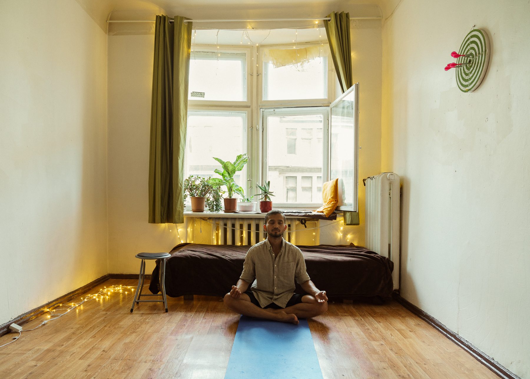 Vīrietis sēž meditācijas pozā uz zila paklājiņa tukšā istabā; erkera logā puķupodi, gar sienu dīvāns un gaismu virtene