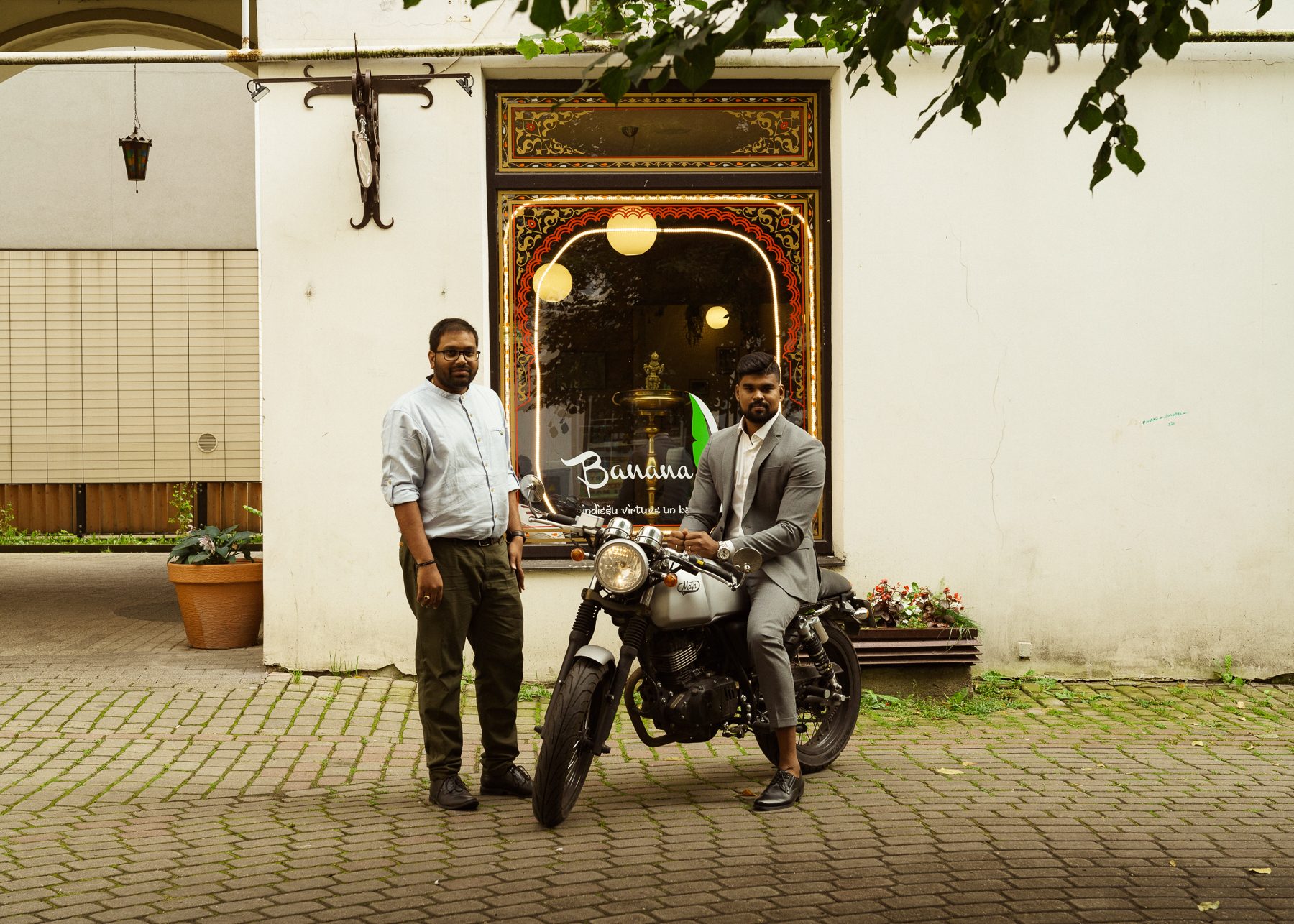 Divi vīrieši pie indiešu restorāna “Banana” rotāta skatloga; viens stāv, otrs pelēkā uzvalkā sēž uz motocikla