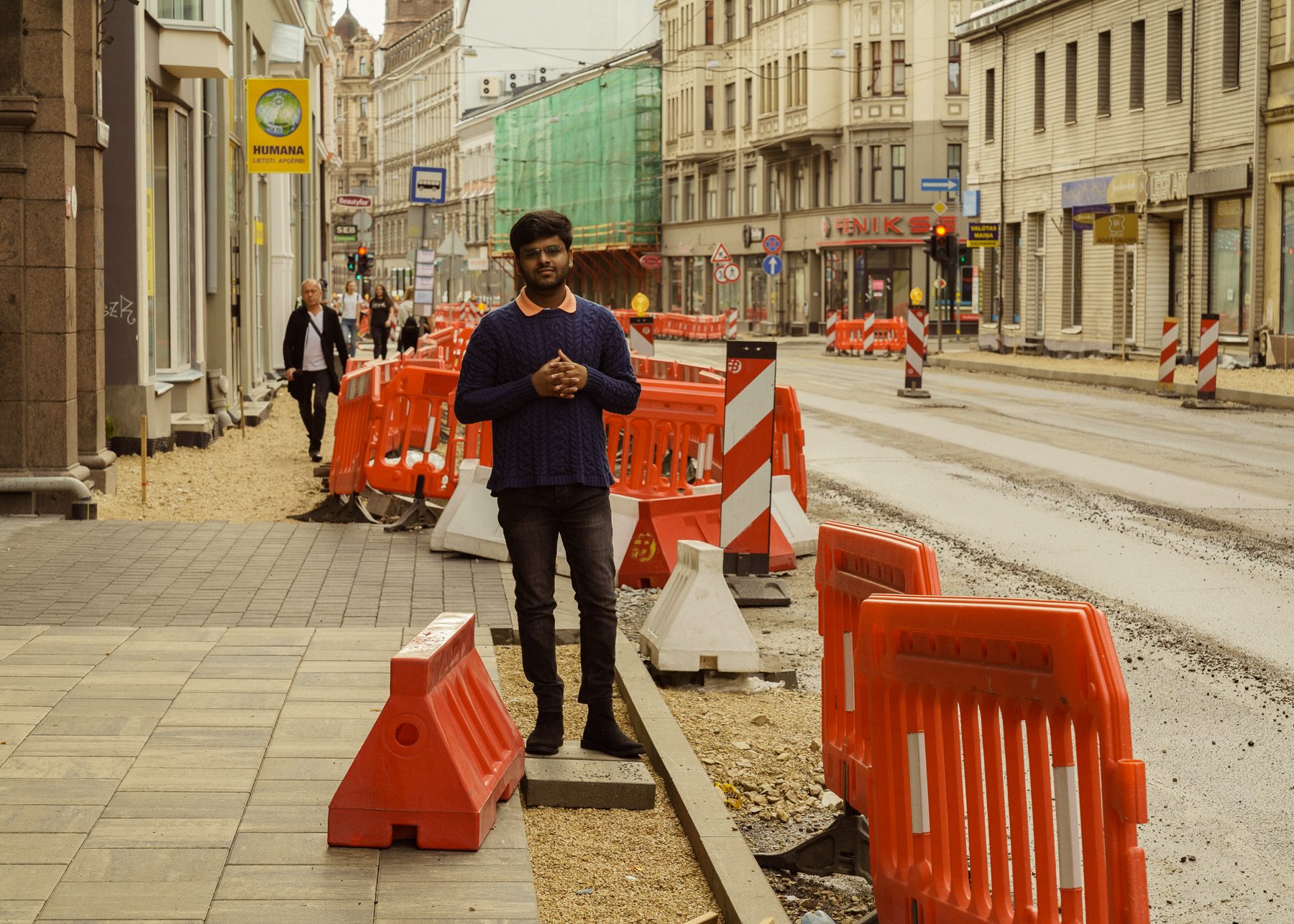Jauns vīrietis tumši zilā džemperī stāv uz uzraktās ietves starp oranžām ceļa barjerām Rīgas ielā remontdarbu laikā