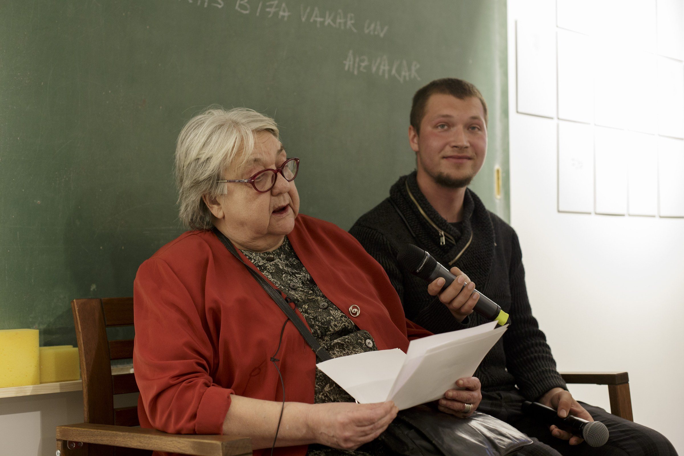 Sieviete sarkanā žaketē sēž un lasa no lapas; blakus vīrietis ar mikrofonu, aiz viņiem ar krītu aprakstīta tāfele