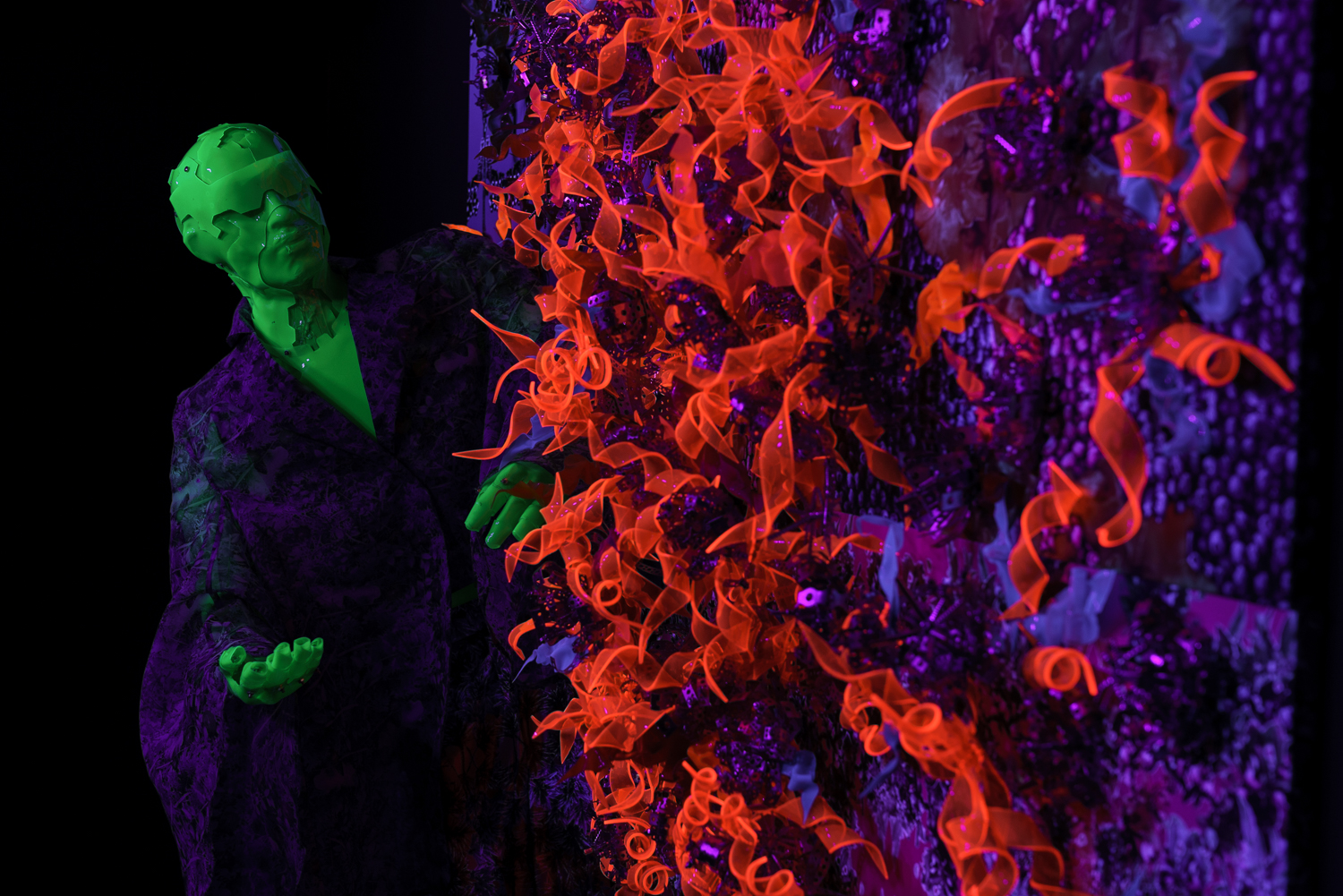 Tuvplānā figūra ar spilgti zaļu seju un rokām pie oranžām fluorescējošām formām ultravioletā gaismā