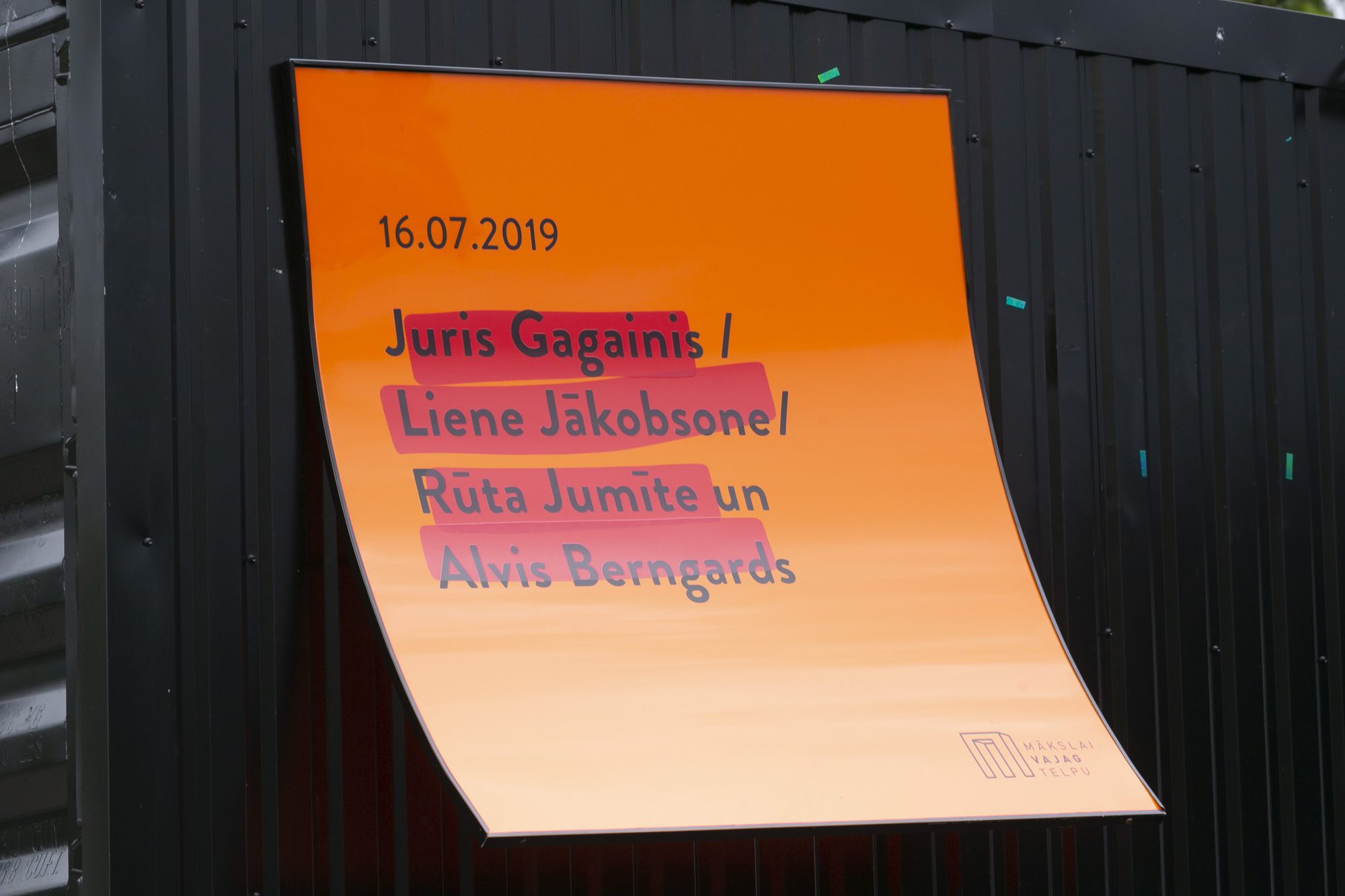 Oranžs izstādes plakāts uz melnas konteinera sienas: „16.07.2019 Juris Gagainis / Liene Jākobsone / Rūta Jumīte un Alvis Berngards”