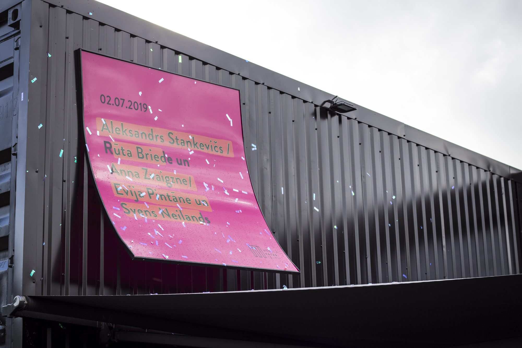 Rozā izstādes plakāts ar datumu 02.07.2019 un mākslinieku vārdiem uz melnas konteinera sienas
