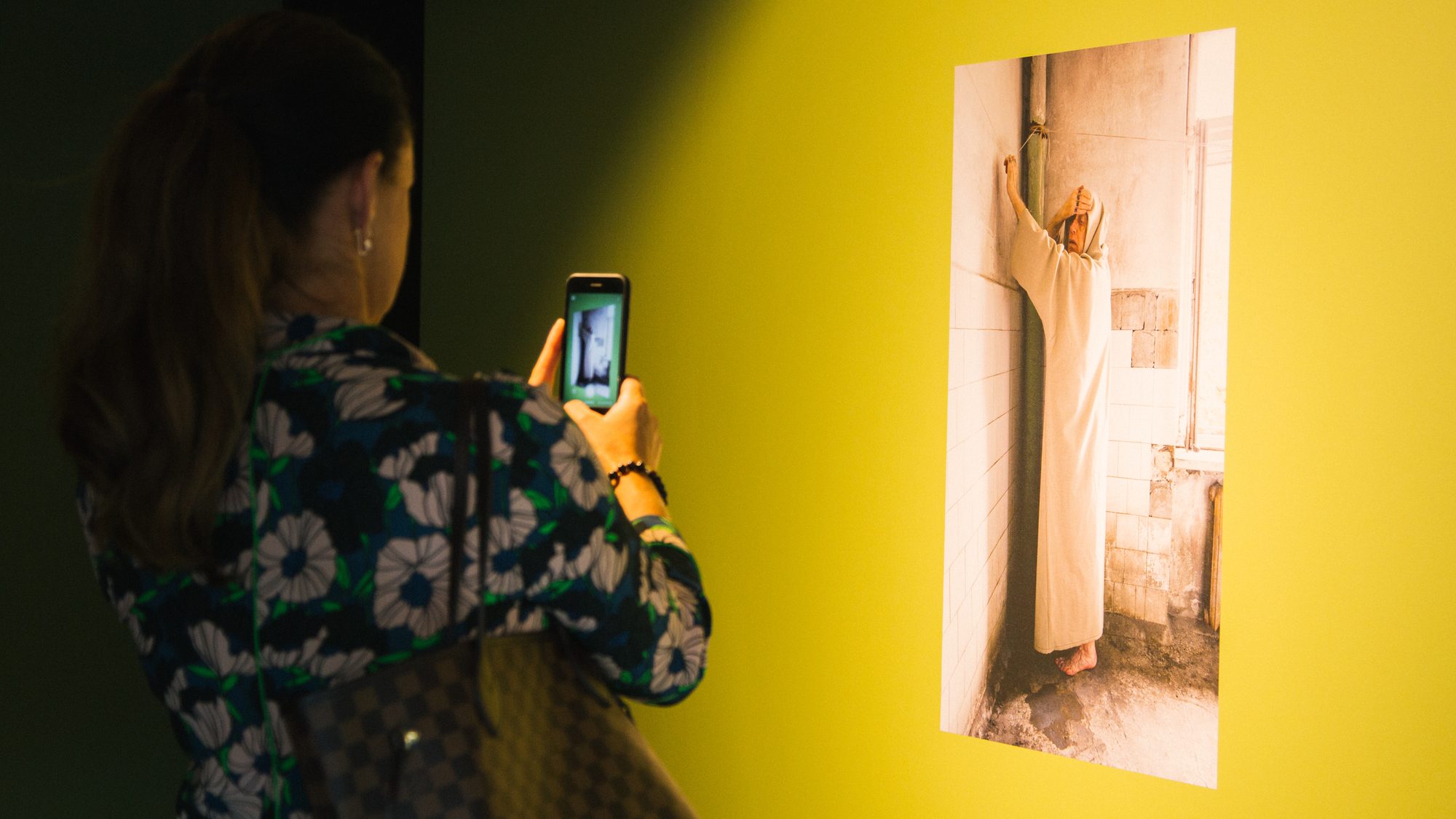 Apmeklētāja fotografē izstādes fotogrāfiju ar figūru garā baltā tērpā