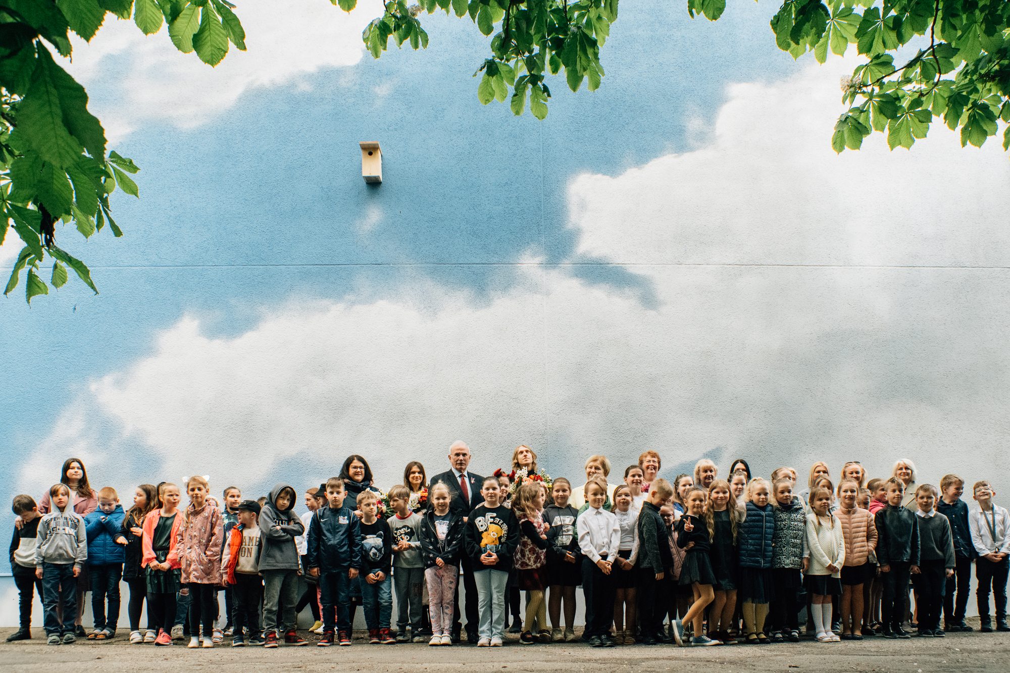 Jāņa Šneidera murālis — ēkas siena, apgleznota kā zilas debesis ar mākoņiem, augšā piestiprināts putnu būrītis; sienas priekšā skolēnu grupa