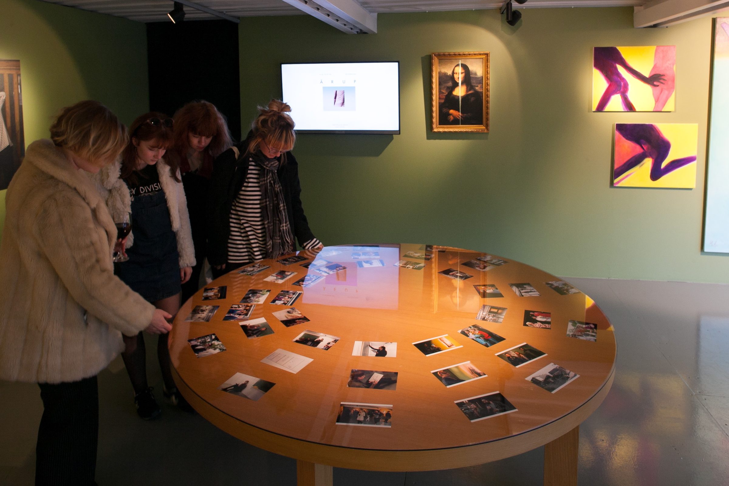 Apmeklētājas pie apaļa galda ar izklātām fotogrāfijām; uz ekrāna uzraksts „ĀRUP”, blakus Monas Lizas reprodukcija un gleznas