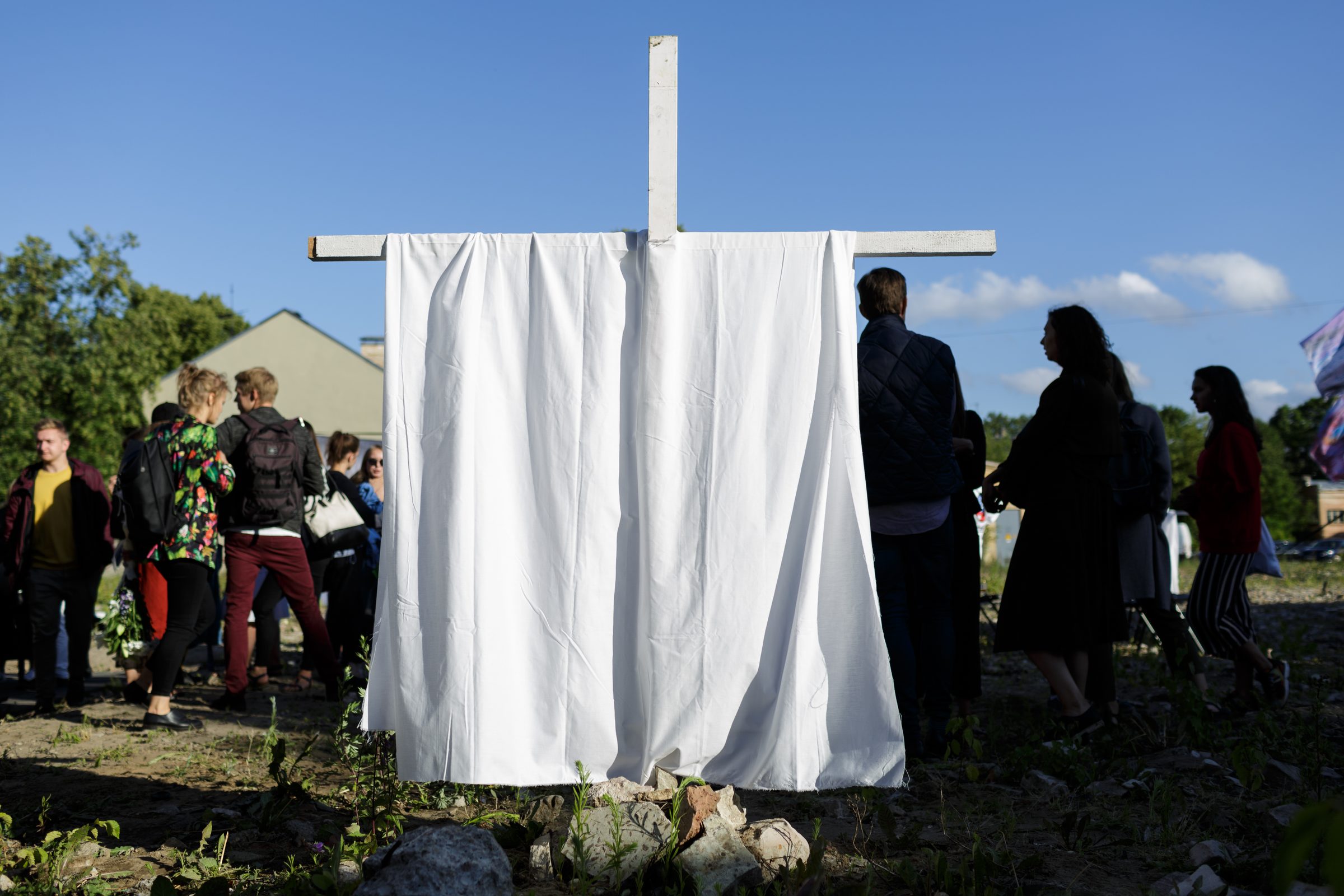 Pagalmā uzstādīts balts krusts ar pārmestu baltu audumu; apkārt apmeklētāji