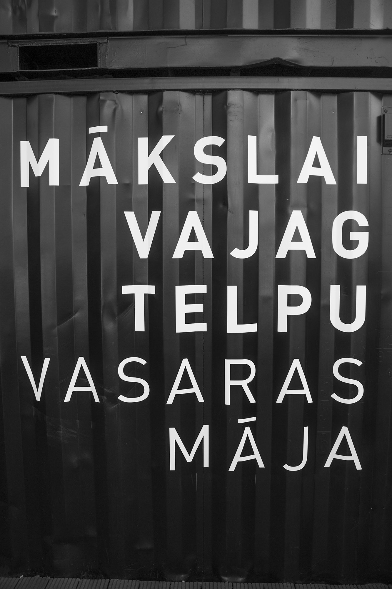 Balts uzraksts „Mākslai vajag telpu. Vasaras māja” uz melna konteinera sienas