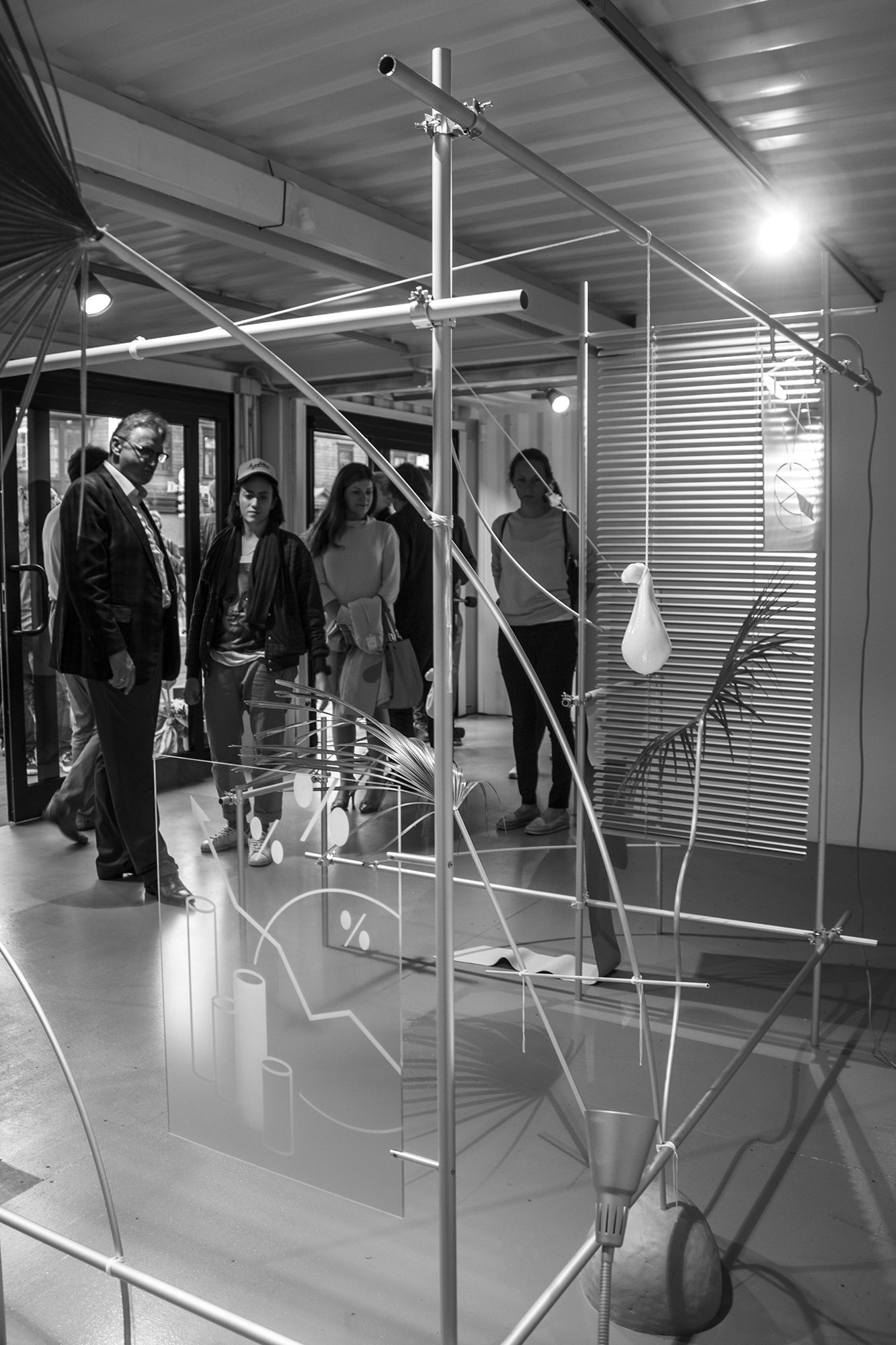 Skats cauri alumīnija cauruļu konstrukcijai: žalūzijas, palmu lapas un siera bloki naudas maisiņu formā; fonā apmeklētāji
