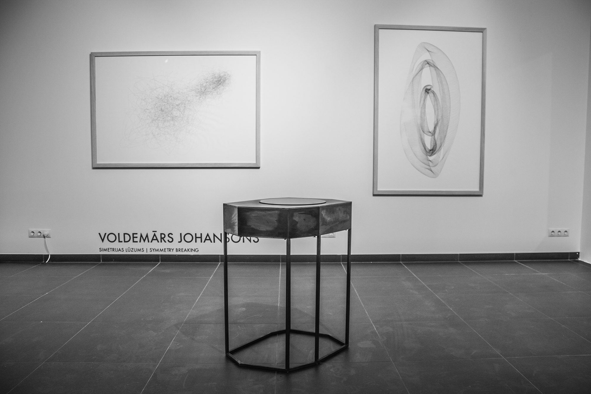 Izstāžu zāle ar diviem ierāmētiem zīmējumiem un objektu uz metāla statīva; pie sienas uzraksts „Voldemārs Johansons. Simetrijas lūzums”