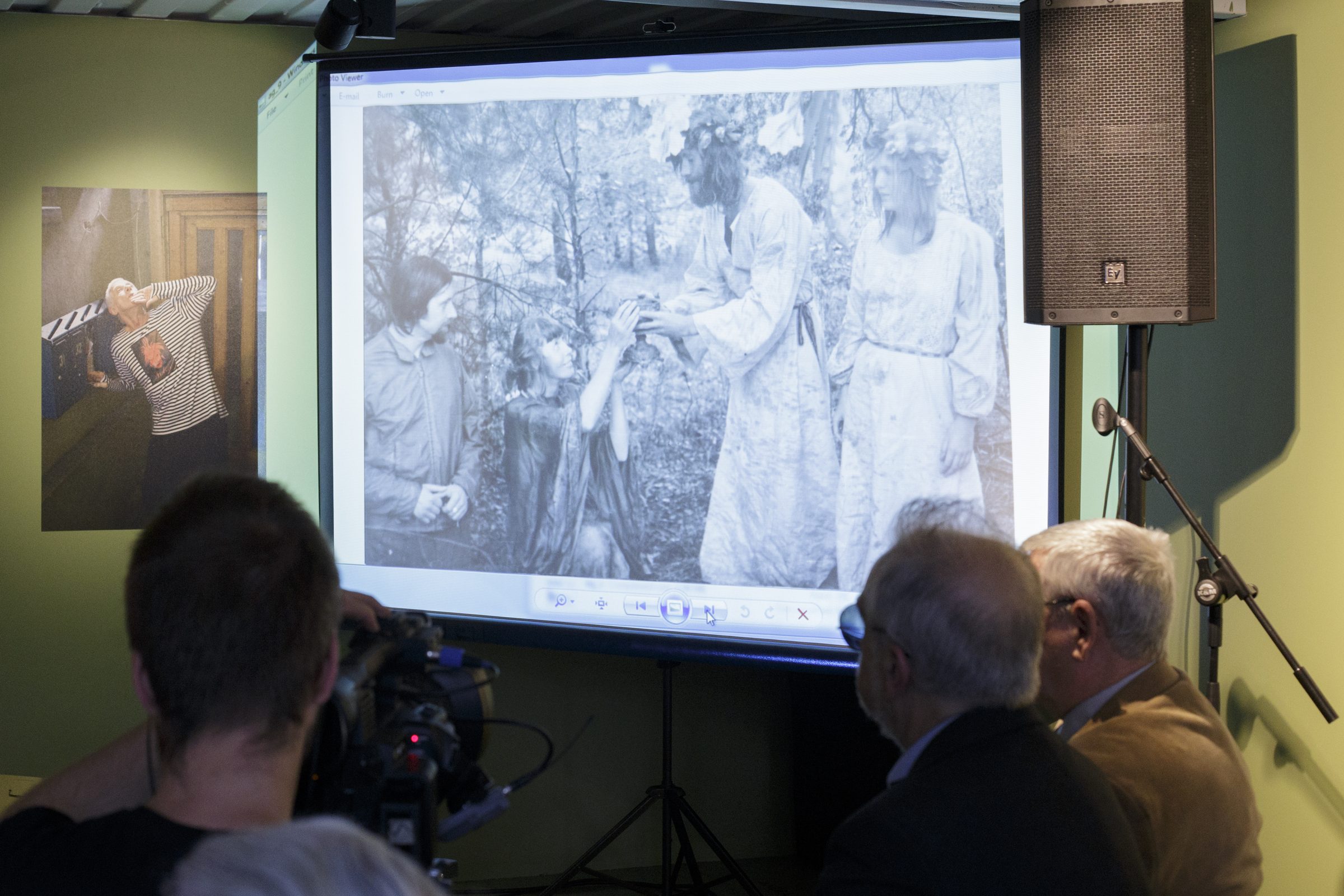 Uz ekrāna projicēta melnbalta fotogrāfija ar cilvēkiem mežā; priekšplānā skatītāji