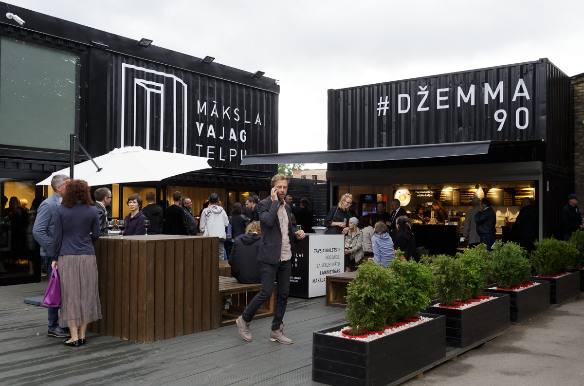MVT Vasaras mājas konteineri ar uzrakstiem „Mākslai vajag telpu” un „#Džemma 90”; priekšā uz koka klāja apmeklētāji