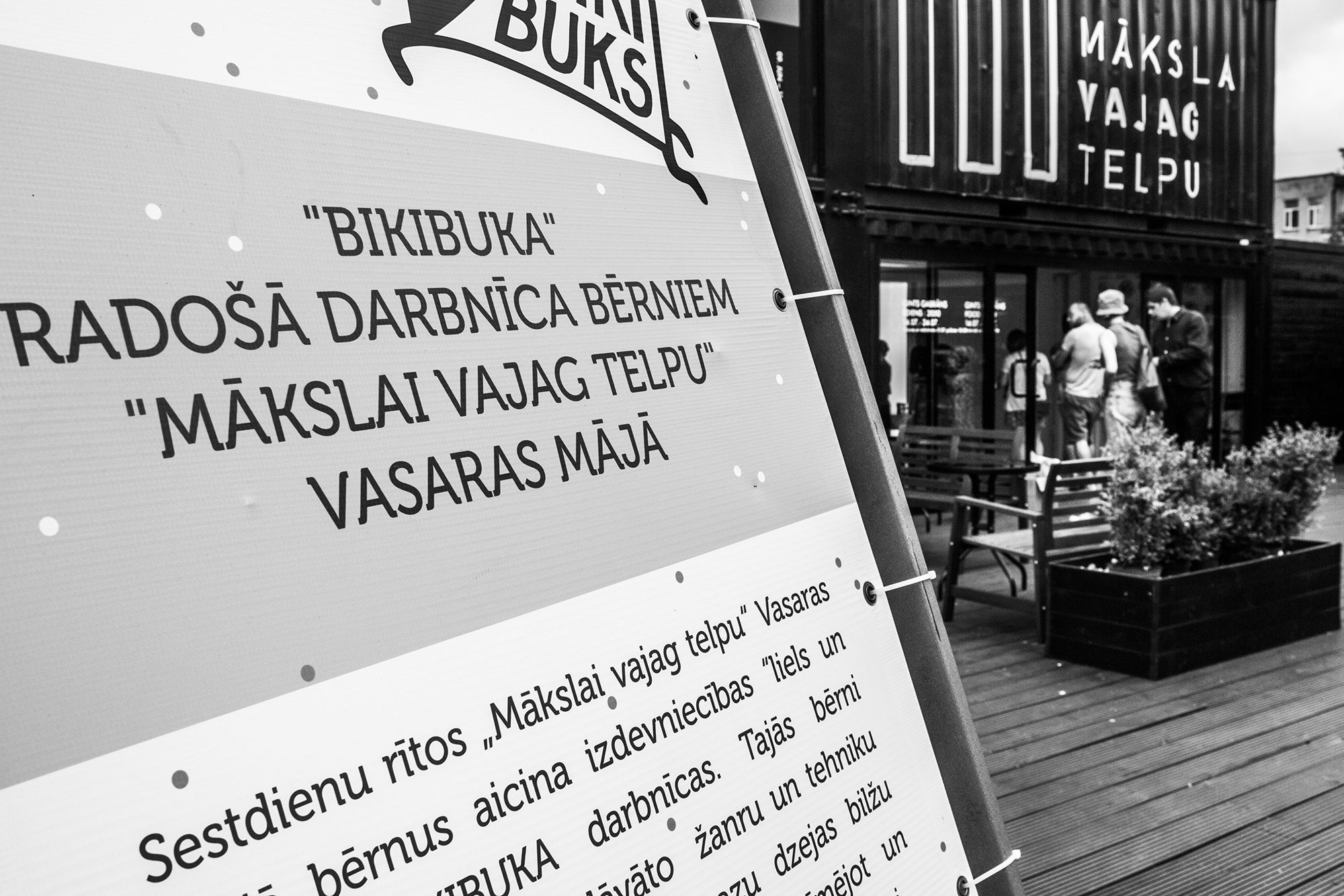 Stends ar uzrakstu „“BIKIBUKA” radošā darbnīca bērniem “Mākslai vajag telpu” Vasaras mājā”; fonā konteiners