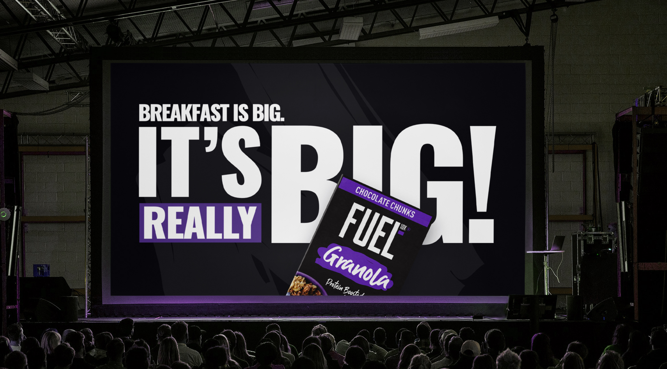 Audience watches a large screen displaying an ad: 'Breakfast is big. It’s really big!' with Fuel Granola pack.
