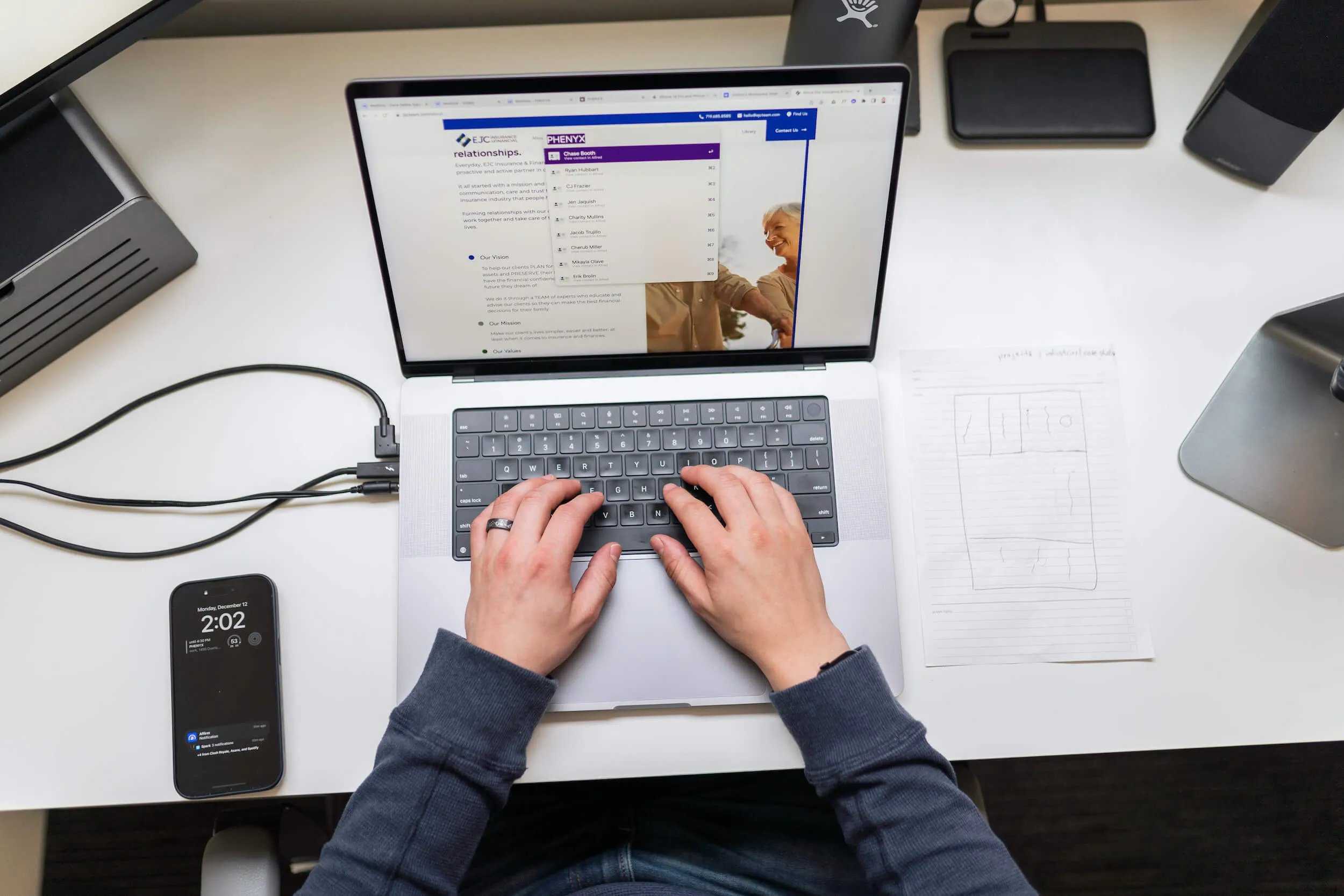 Overhead view of hands typing on a laptop showing a client insurance website, beside a phone and a hand-drawn page wireframe