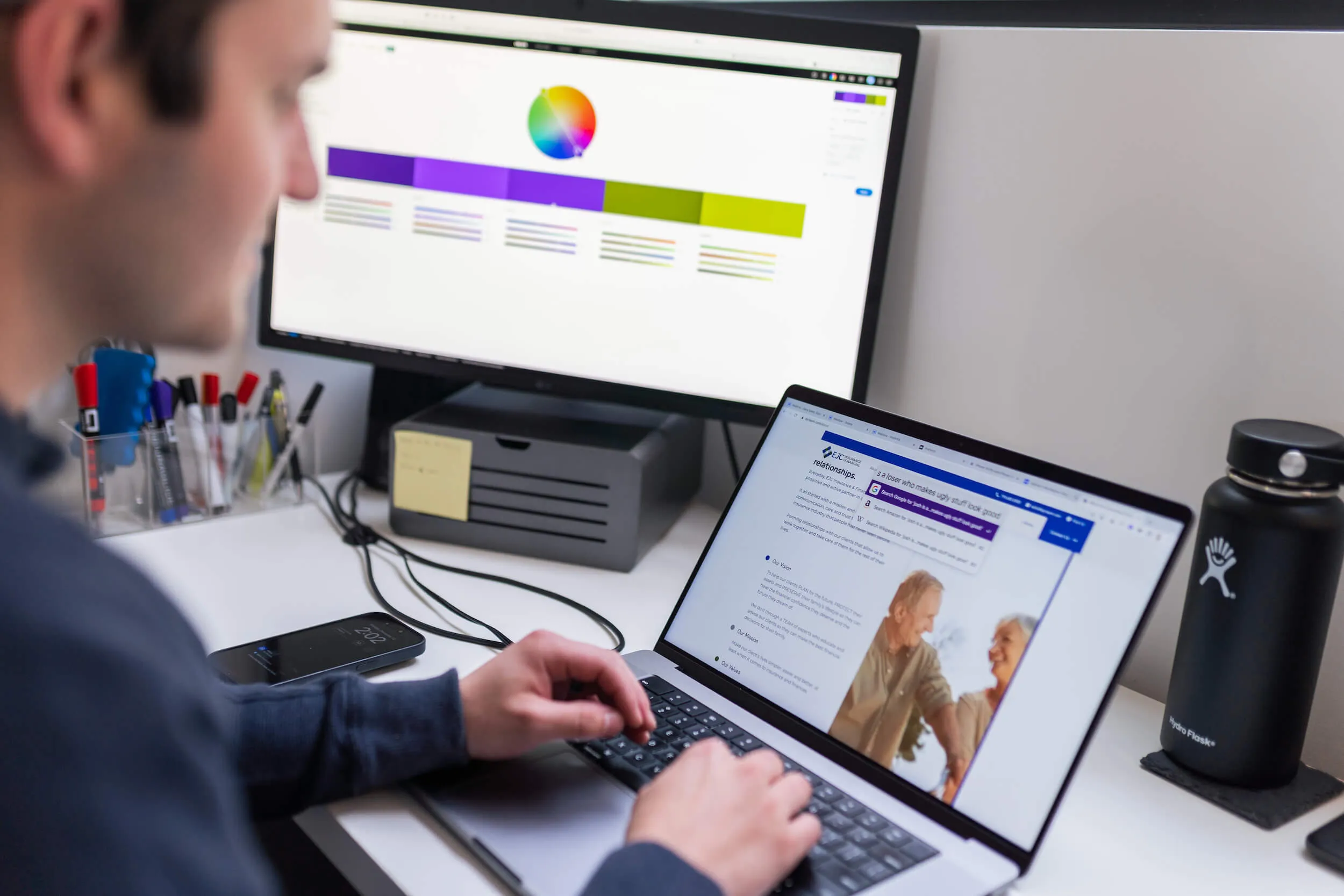 A PHENYX designer reviewing a brand colour palette on a monitor with a client website open on the laptop beside it