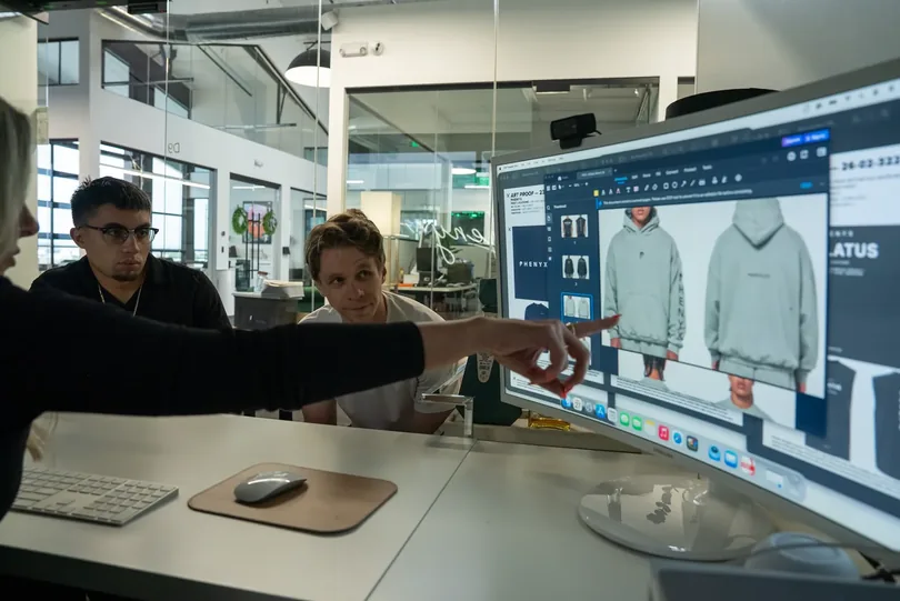 Three PHENYX team members reviewing apparel mockups on a large studio monitor, one pointing at the screen