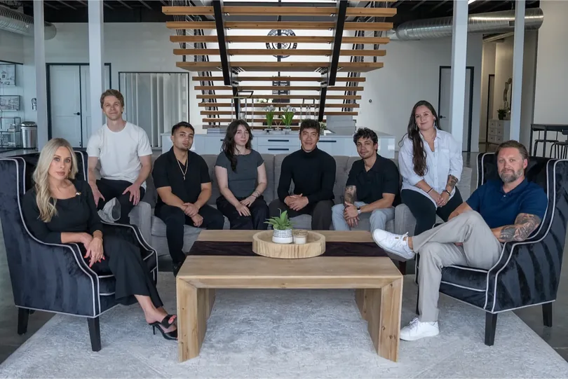 The full PHENYX team of eight seated together in the studio lounge beneath a slatted wood ceiling